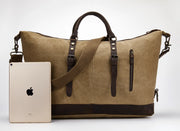 A large tan canvas duffel bag with dark brown leather handles, straps, and trim, shown on a light gray studio background next to a silver iPad for scale.