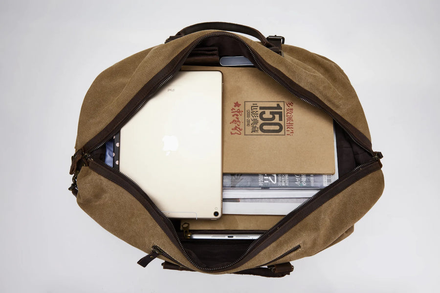 A top-down view of a brown canvas duffel bag with its main compartment open, showing a white laptop and a brown paper box inside.
