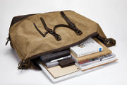 A tan canvas duffel bag with dark brown leather straps and buckles, shown open with its contents spilled out on a white studio background. Visible items inside include books, a tablet, a notebook, a pen, and a wallet.
